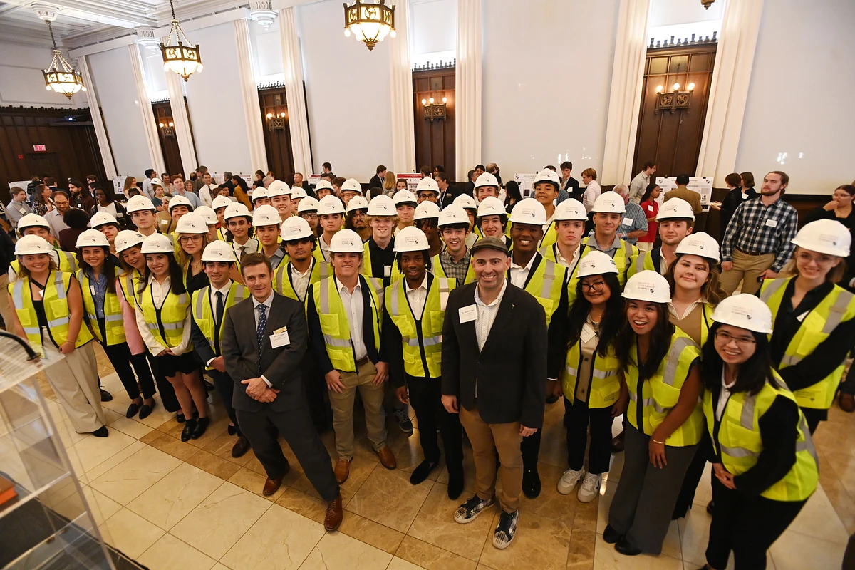 A large group of people wearing hard hats and yellow safety vests pose for a photo in a spacious, chandelier-lit room.